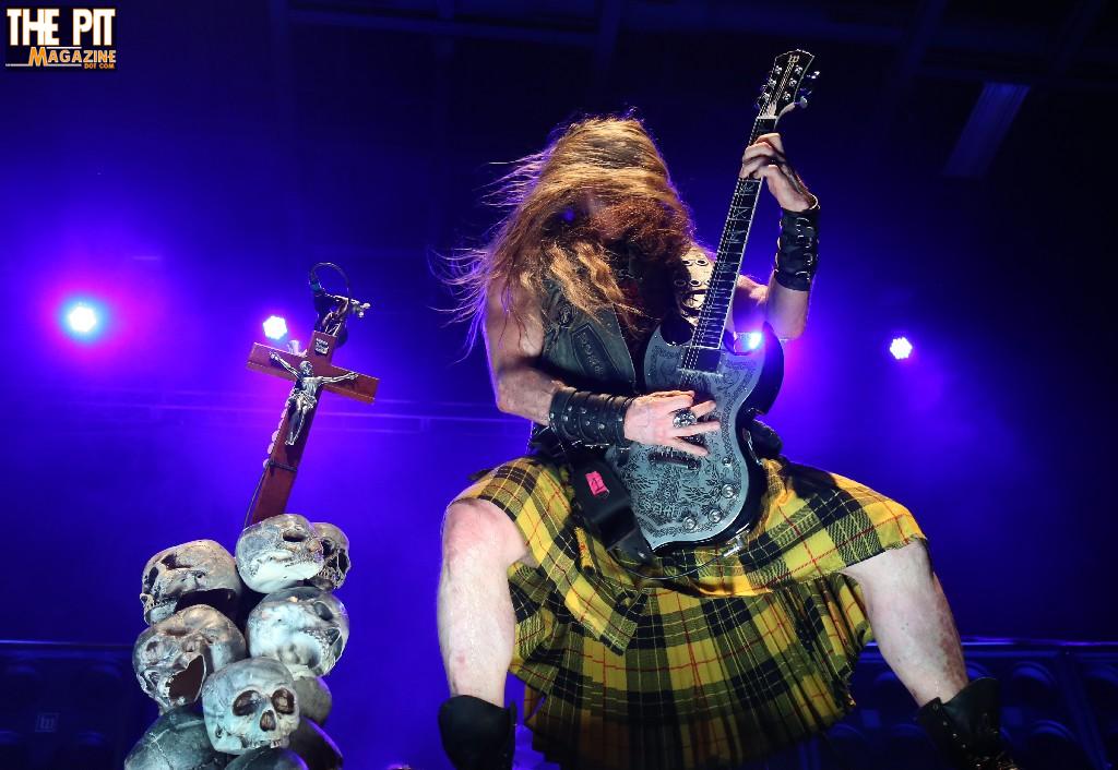 Guitarist with long hair and kilt, performing energetically on stage, surrounded by Black Label Society skull decoration under blue lighting.