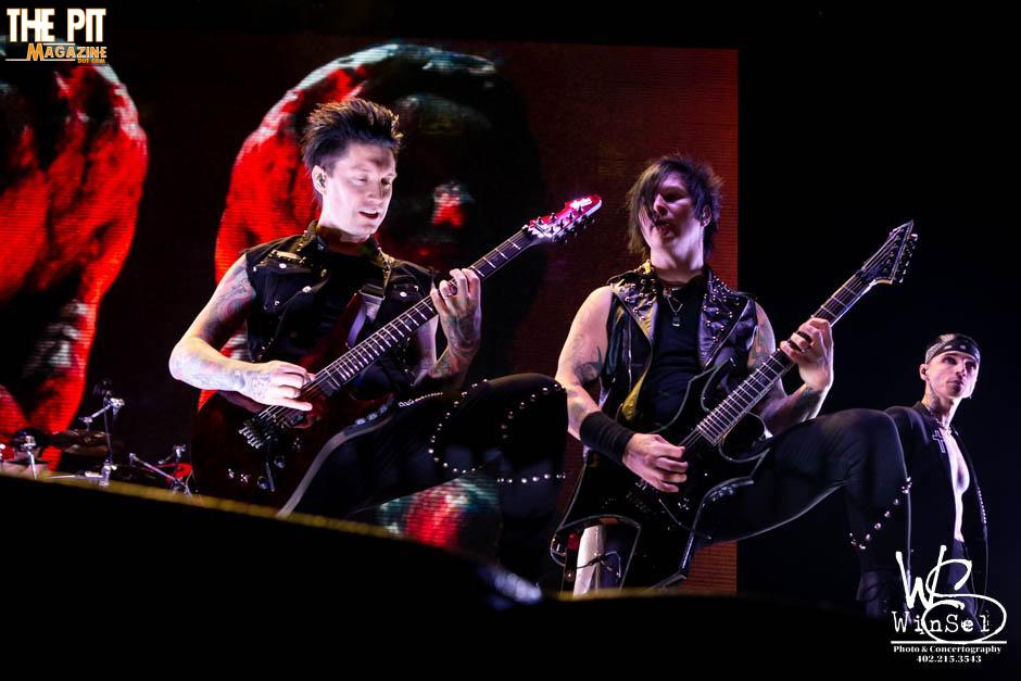 Two guitarists and a drummer from Black Veil Brides perform energetically on stage, surrounded by dramatic stage lighting and a large screen displaying red visuals.
