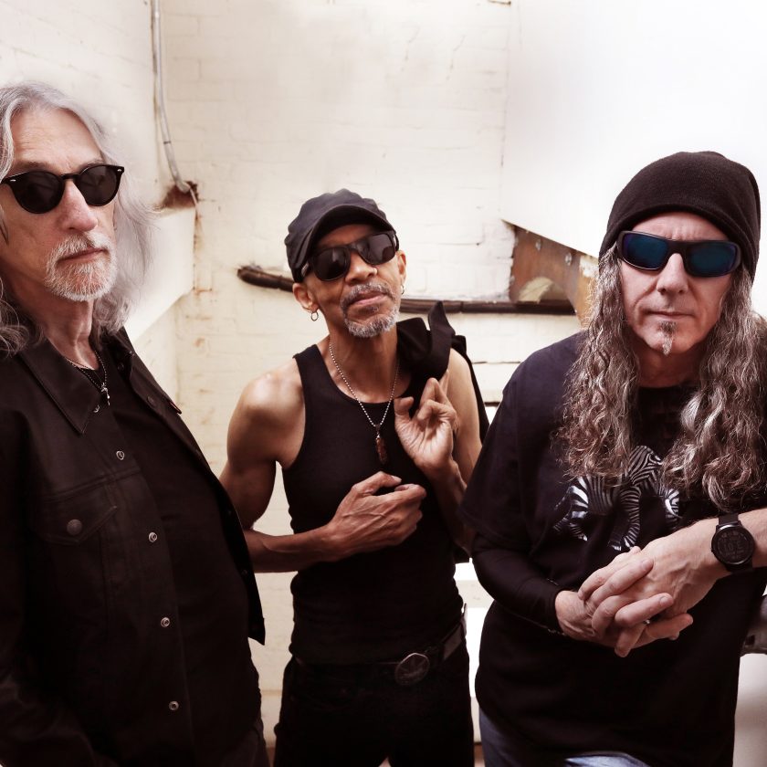 Three middle-aged male musicians standing together in a room with white walls, one wearing sunglasses and a black cap, another in aviator shades, and the third in a King's X band t-shirt.