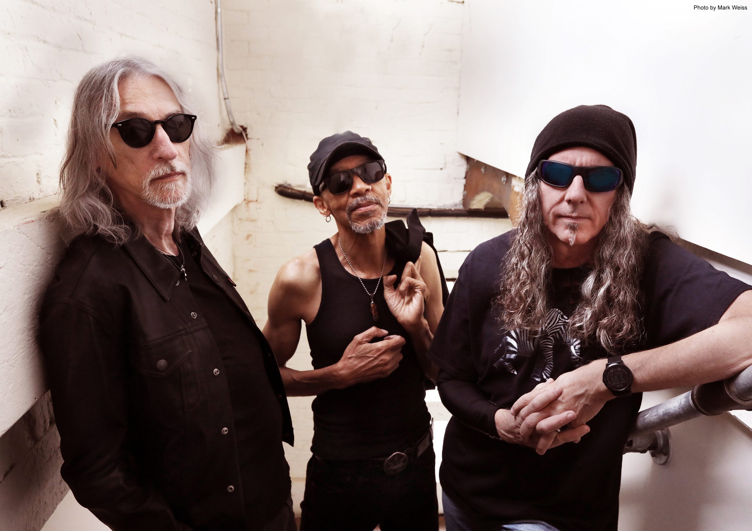 Three middle-aged male musicians standing together in a room with white walls, one wearing sunglasses and a black cap, another in aviator shades, and the third in a King's X band t-shirt.