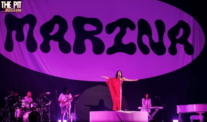 A female singer in a red outfit performing on stage, with a large sign reading "Marina" in the background and band members playing instruments.