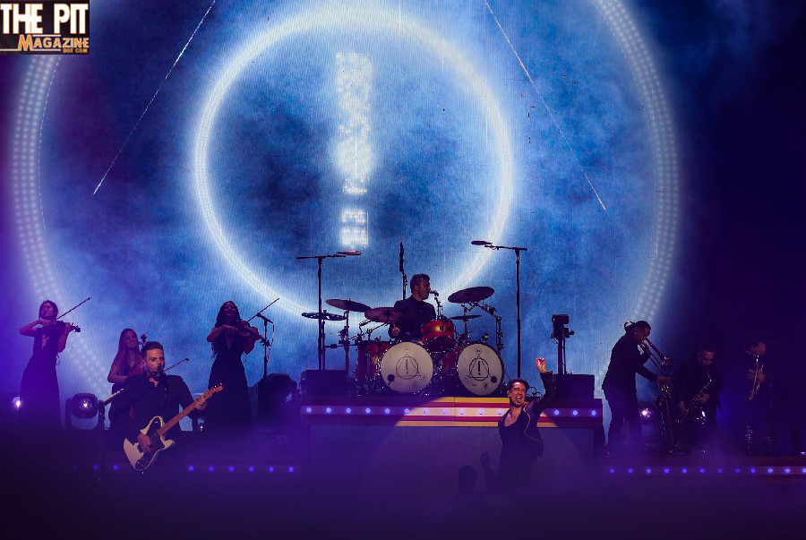 Panic! At The Disco performing on stage under a large illuminated moon graphic, with the lead singer in the foreground and musicians with various instruments in the background.