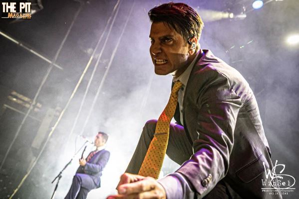 A man in a suit and tie passionately performing on stage with a microphone, intense lighting, and smoke around him; another performer from Ice Nine Kills is in the background.