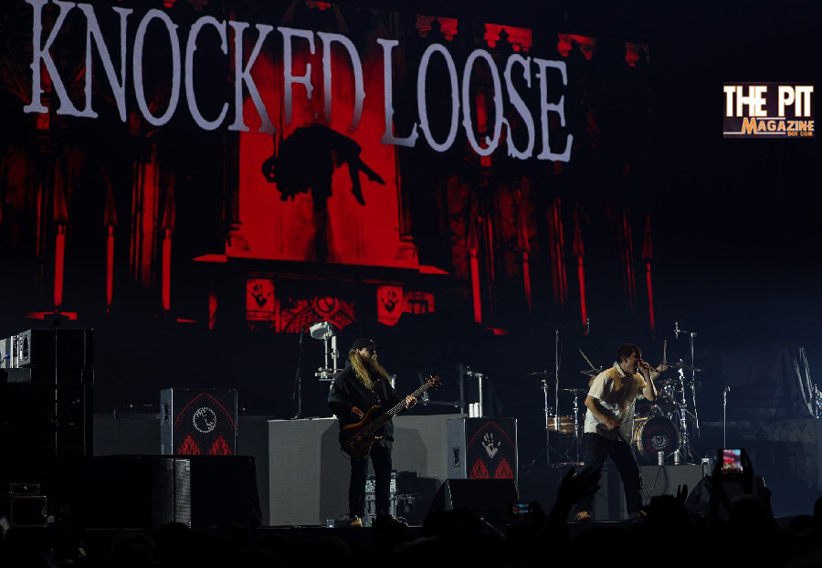 Band members of Knocked Loose perform on stage under red lighting with their logo displayed on a large banner behind them.