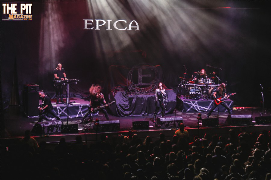 Concert scene with band Epica performing on stage, showing six members playing instruments and singing under purple stage lights, with the band's logo projected in the background.