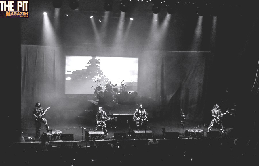 Black and white photo of metal band Sabaton performing on stage, with members playing guitars and drums under dramatic lighting.