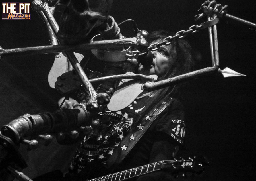 Black and white image of two WASP rock musicians playing guitar and bass closely together on stage, both wearing elaborate costumes and sunglasses.