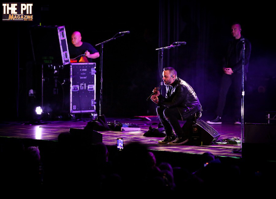 A male singer in a black leather jacket kneels on stage, performing into a microphone, with Blue October band members in the background under purple stage lighting.
