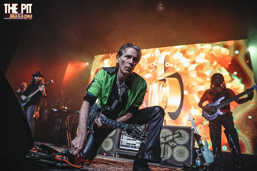 Lead singer in a green shirt and black jeans crouching on stage, with band members including Steve Vai playing guitars in the background, bright stage lights and vibrant screen.
