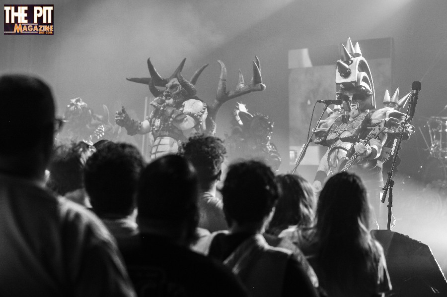 GWAR band members in elaborate costumes perform on stage in front of an audience at a concert, captured in black and white.