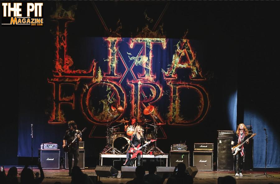 Rock band performs on stage under a large "Lita Ford" logo, with a drummer and two guitarists in front of amplifiers.