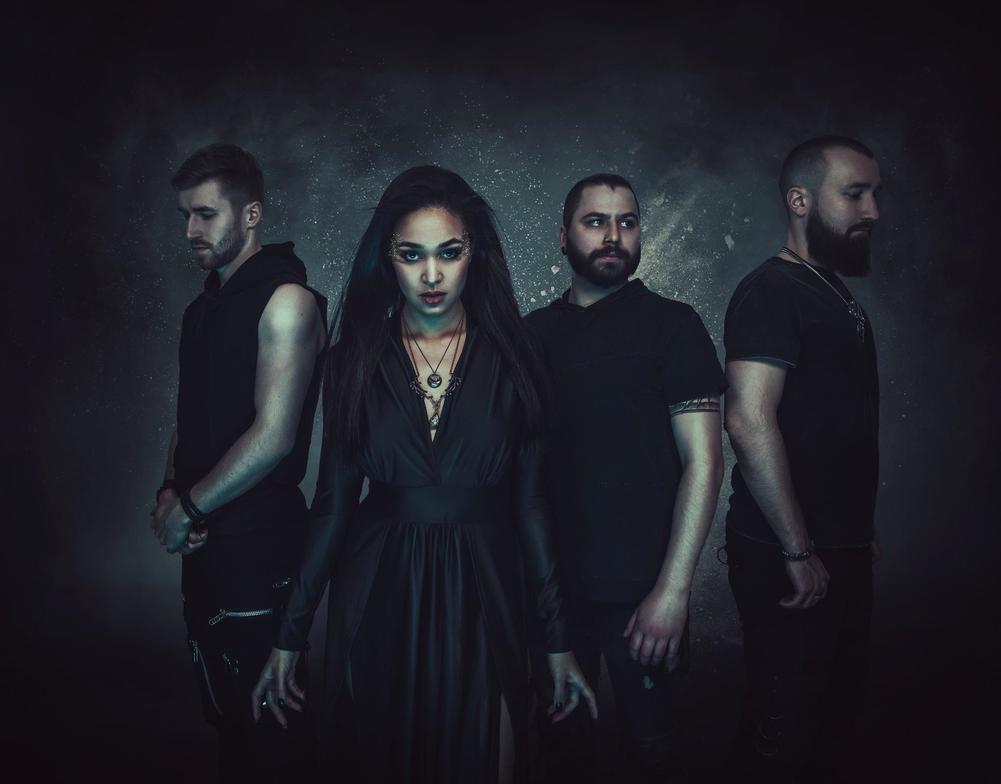 Four people, three men and one woman, ad infinitum stand in a dark, moody setting, dressed in black, with a focus on the woman in the center.