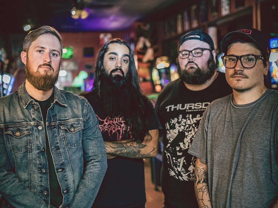 Four men standing in a bar with colorful lighting in the background, looking at the camera, three with beards, one in an American Standards denim jacket.