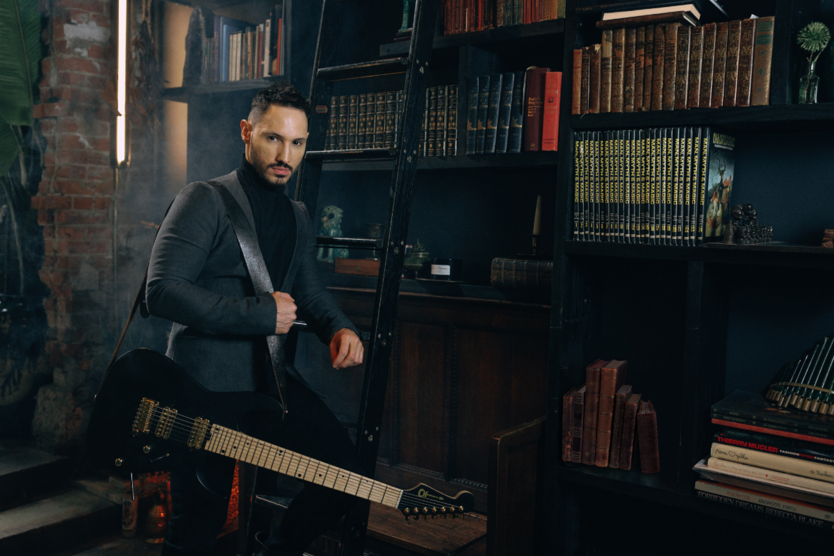 Angel Vivaldi, holding an electric guitar, sits on a stool in a room filled with bookshelves and dim lighting.