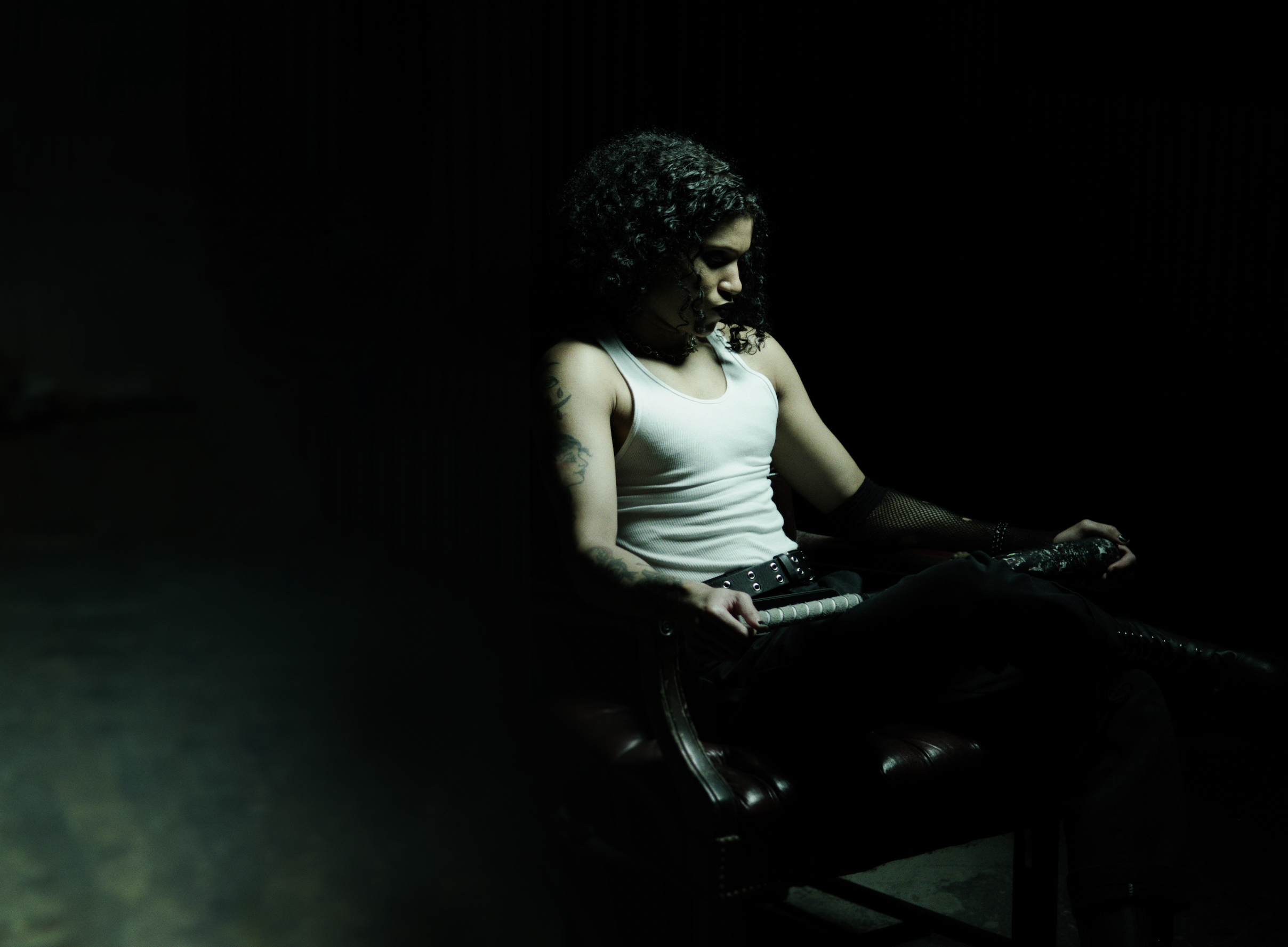 A person with curly hair and tattoos sits on a leather chair in a dimly lit room, contemplatively looking downward, resembling a scene from a Bloodbather music video.