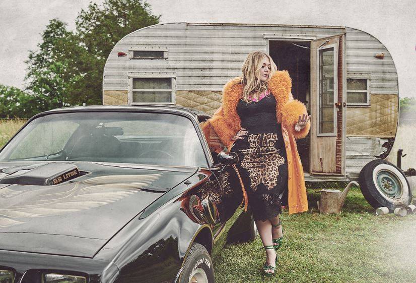 Elle King in a vibrant outfit standing beside a classic car near an old trailer in a rural setting.