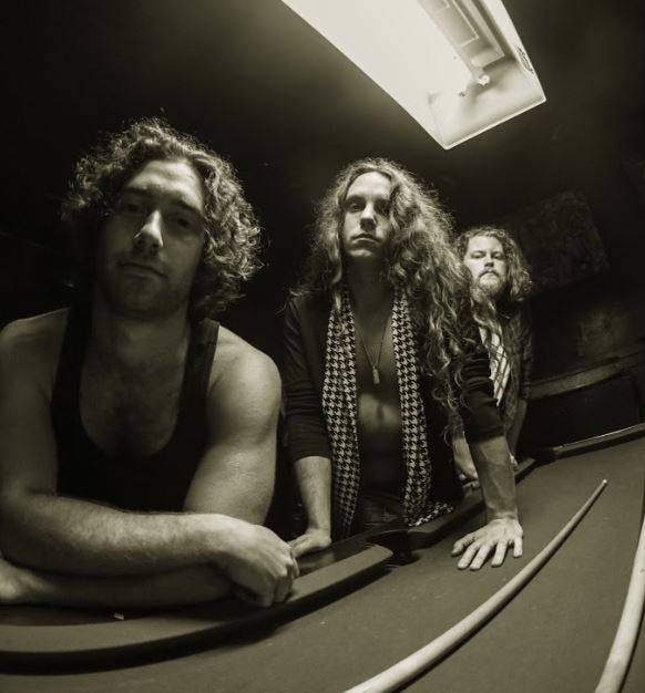 Three men with long hair posing seriously around a curved counter, illuminated by an overhead light in a dimly lit room, resembling members of Fortune Child.