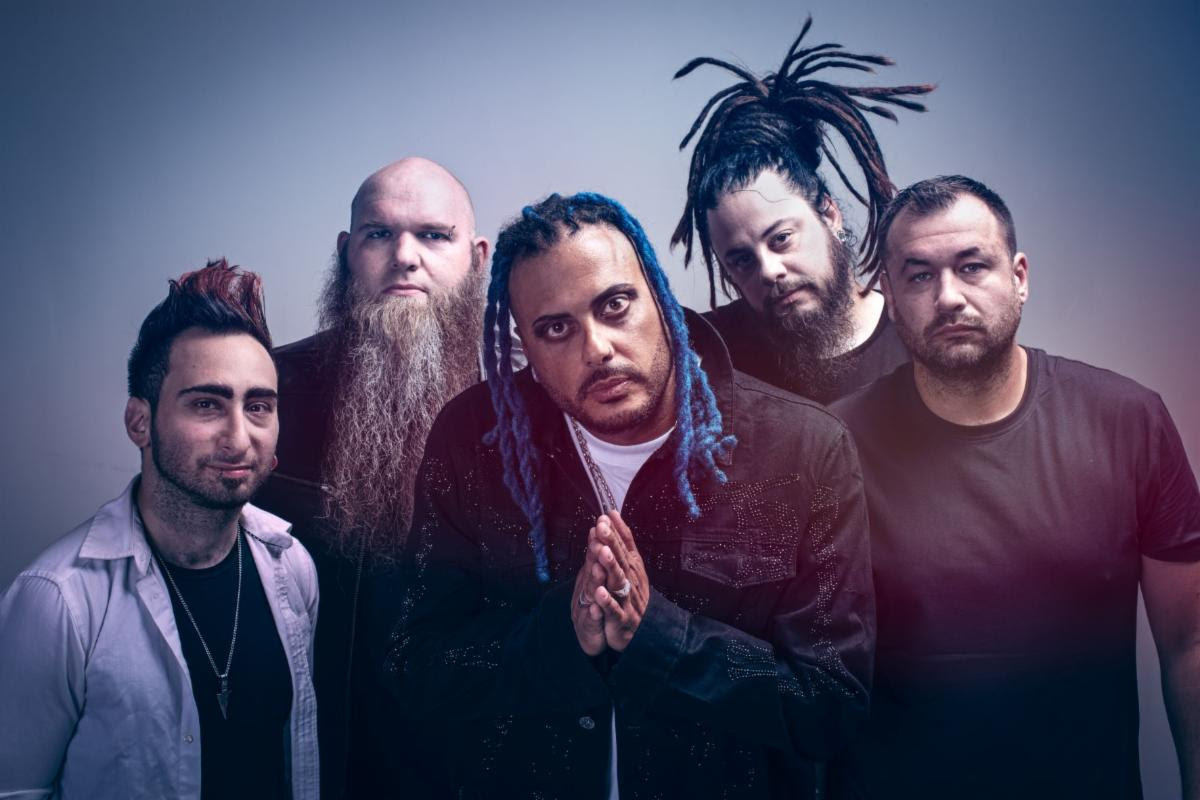 Five men posing for a group photo against a backdrop adorned with gears, with diverse hairstyles including braids and a mohawk, looking intently at the camera.