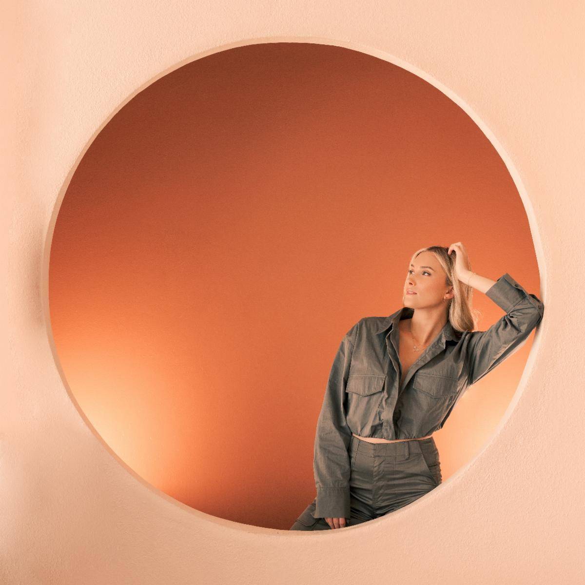A woman in a gray outfit poses thoughtfully within a large circular orange frame against a matching orange background. Juliana Tucker stands confidently, her expression contemplative.