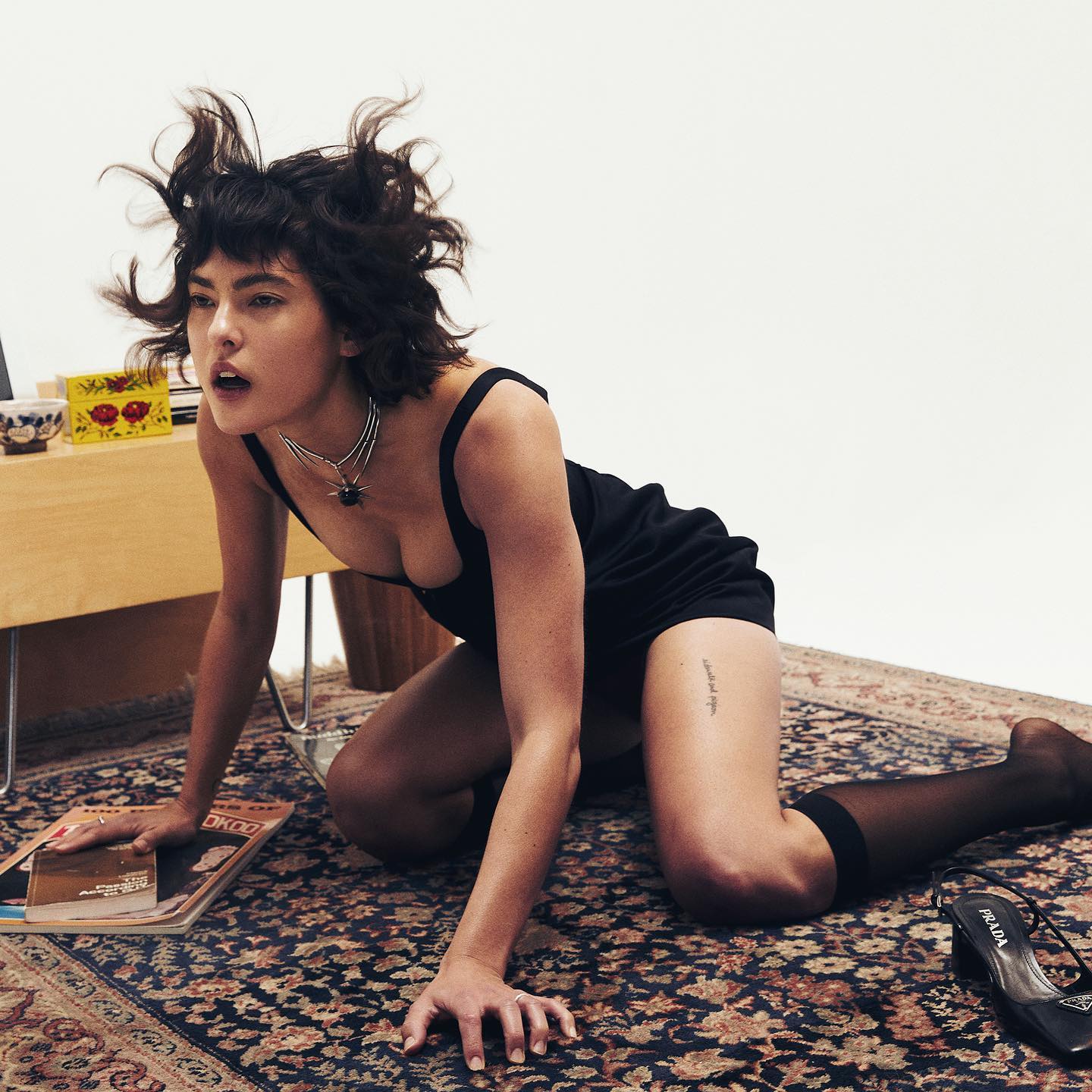 Miya Folick in a black dress crawling on a carpet, looking intensely at the camera with her hair blowing upwards.