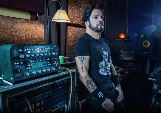 A man with tattoos sits by prong audio equipment in a dimly lit studio, looking pensively to the side.