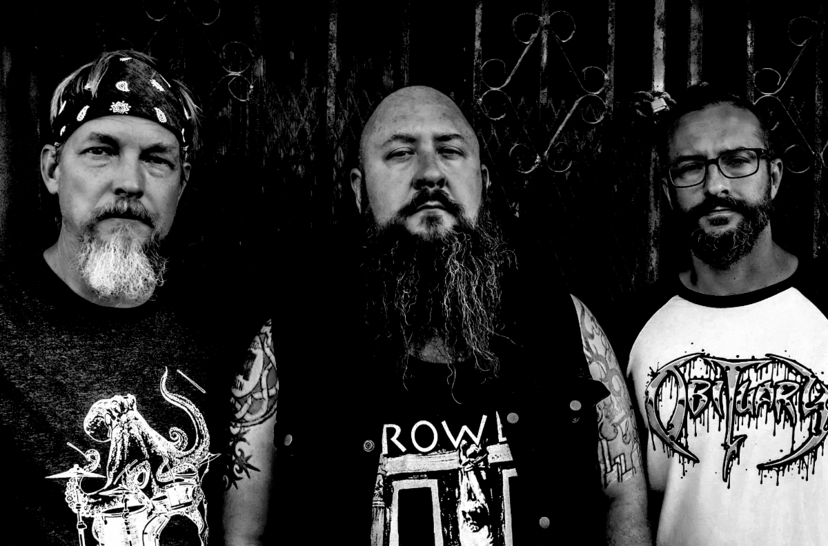 Three men standing against a metal gate, with the middle man having a long beard and the other two sporting shorter facial hair, all wearing Stone Nomads t-shirts.