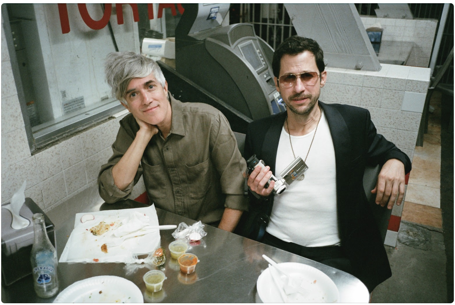 Two members of We Are Scientists sitting at a diner table with empty food containers, one holding a camera. One has grey hair, the other wears a black jacket and sunglasses.