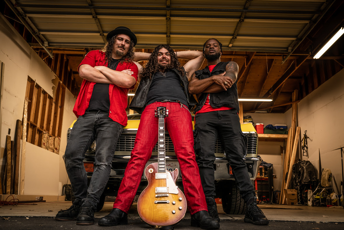 Three musicians from Zach Waters Band in a garage, one holding a guitar, all posing confidently with arms crossed or on hips.