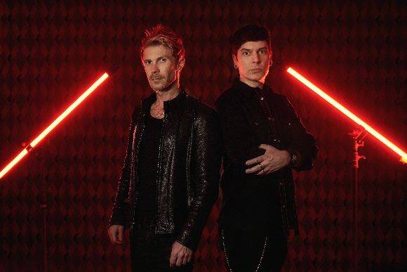 Two men in black outfits stand against a red patterned background with diagonal d3adc0de light beams.