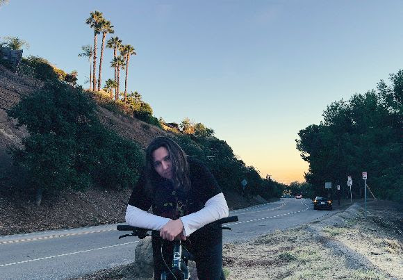 A person with long hair leaning on a bike beside a roadside with tall palm trees on a hill and cars in the distance at sunset, listening to Good Bison.