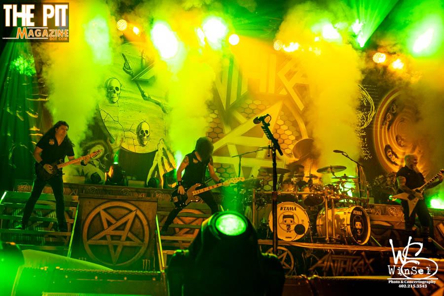 Anthrax performs on stage illuminated by green lights with a backdrop displaying symbols and a skull, in front of a crowd.