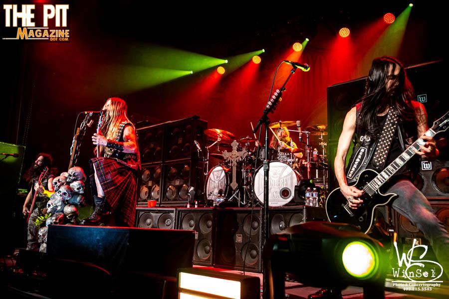A heavy metal band, Black Label Society, performs on stage with vibrant green and red lighting, featuring a guitarist, bassist, and drummer.