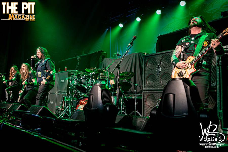 Rock band Exodus performing on stage, with four members playing guitars and drums under blue and green lights, with a logo banner in the background.