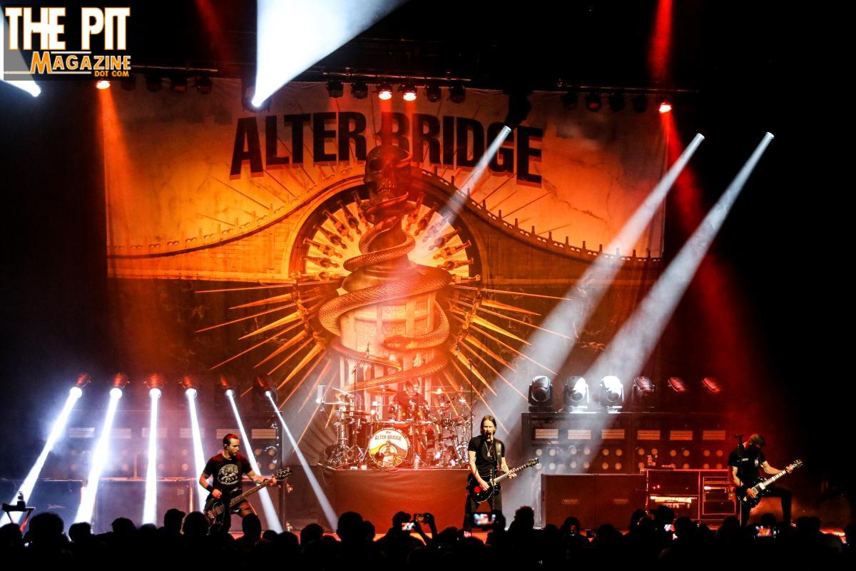 Band members of Alter Bridge perform on stage with dramatic lighting and a large backdrop featuring their logo.