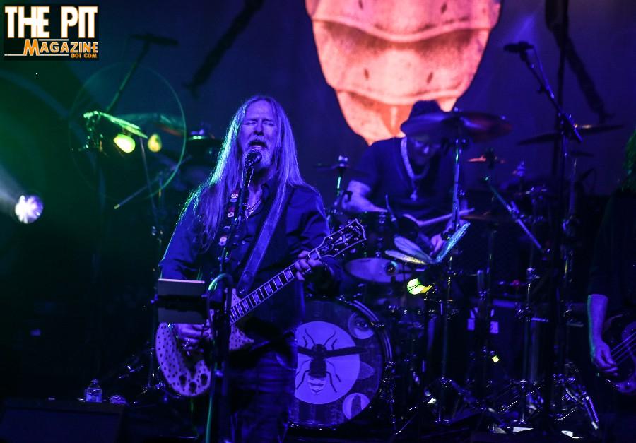 Jerry Cantrell, a guitarist with long blond hair, performs onstage under blue lighting, accompanied by a drummer in the background.