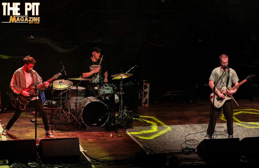 Three band members performing on stage, with a guitarist, drummer, and bassist highlighted under stage lights, reminiscent of a Dead Poet Society performance.