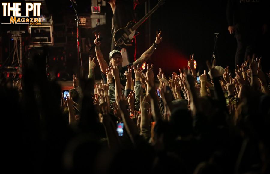 Crowd of Highly Suspect fans cheering and taking photos at a concert, with the performer visible in the background on stage.