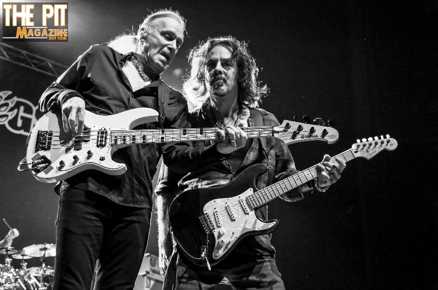 Two male guitarists from Winery Dogs playing electric guitars on stage, with one showing the other his guitar fretboard; black and white photo with “the pit magazine” watermark.