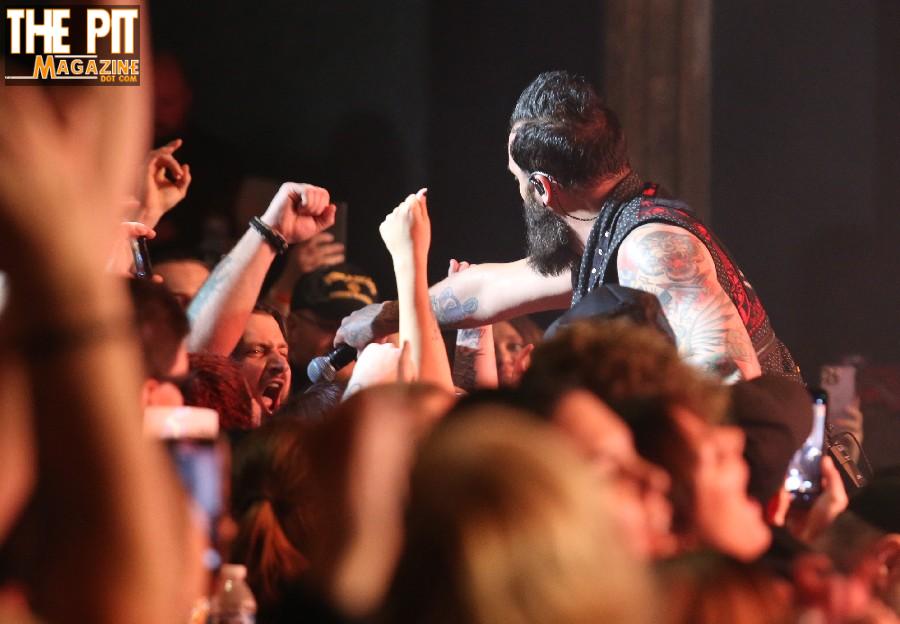 A tattooed singer with a beard from Skillet engaging the audience at a concert, as fans raise their fists and cheer.