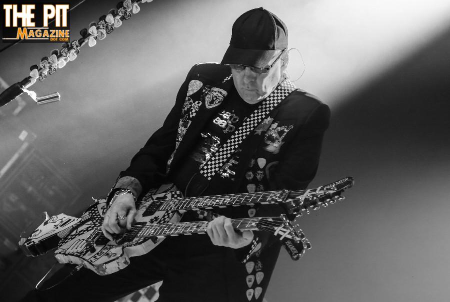 A guitarist in a cap and adorned jacket plays a double-neck guitar on a smoke-filled stage, with the Cheap Trick logo at the top.