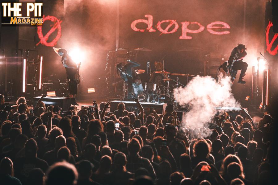 Rock band performing on stage with energetic lighting and a crowd watching, under a sign that reads "Dope.