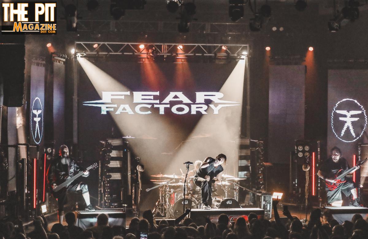 A live concert scene of Fear Factory performing on stage with dramatic lighting, featuring band members with instruments in front of an audience.