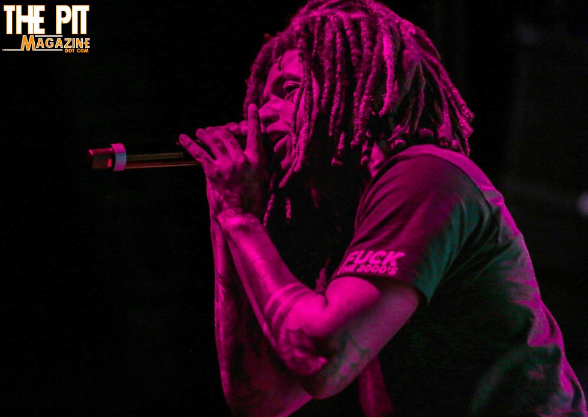 A musician with dreadlocks performs into a microphone on stage, illuminated by red lighting, with the text "City Morgue" visible in the corner.