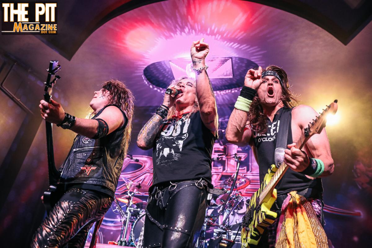 Three Steel Panther musicians performing energetically on stage with a colorful lighting background and the audience's hands visible in the foreground.