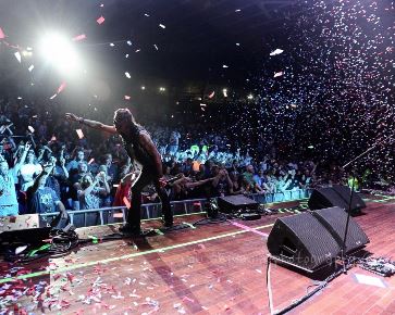A rock musician performing on stage, gesturing to a cheering crowd in a confetti-filled concert hall.