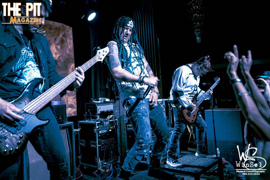 Rock band Above Snakes performing live, featuring a guitarist with long braided hair and a bandanna, engaging enthusiastically with fans. Cool blue stage lighting enhances the intensity of the show.