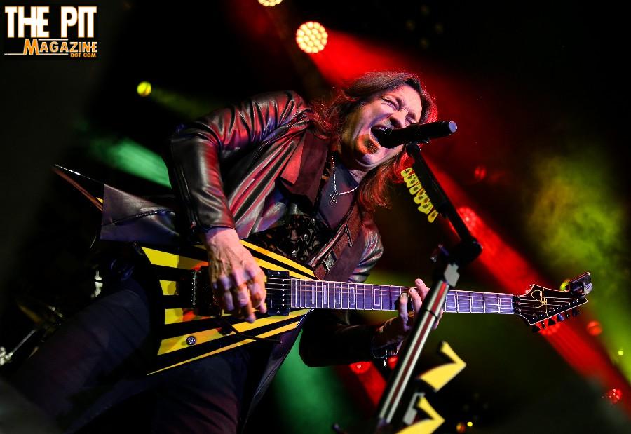 A male guitarist passionately performing on stage under colorful lights, holding a yellow and black Stryper electric guitar.