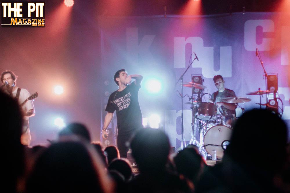 Band performing on stage, lead singer with microphone, audience in foreground, vibrant stage lighting, "Knuckle Puck" logo visible in top left.