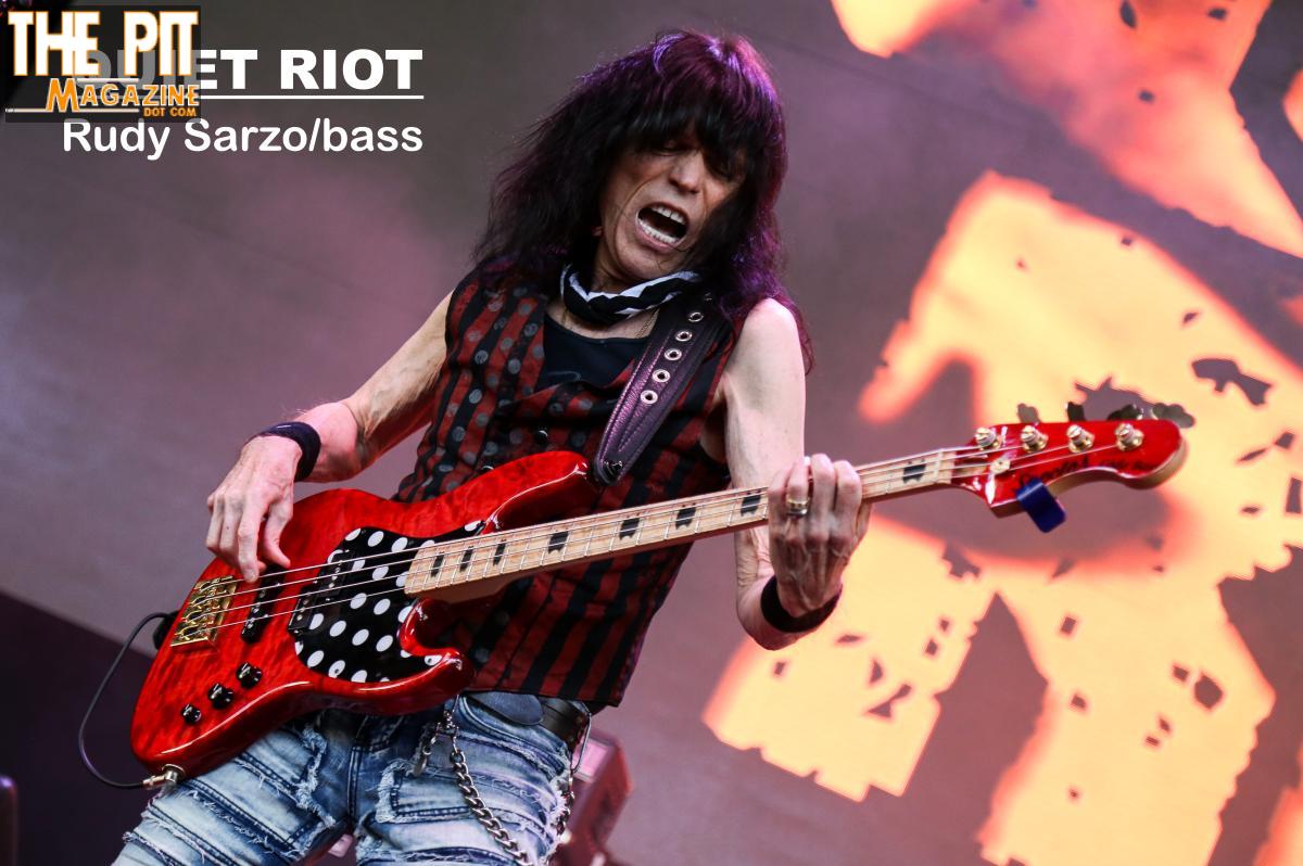 Bassist performing energetically at the M3 Rock Festival, wearing a red and black outfit and playing a red electric bass.