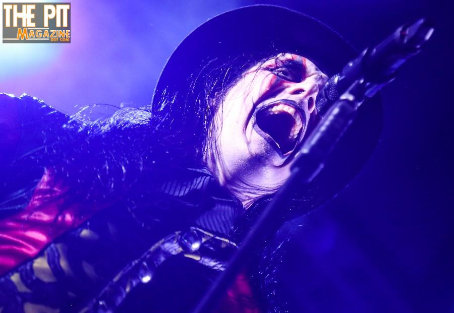 Avatar rock musician wearing a hat and leather, singing passionately into a microphone on a blue-lit stage.